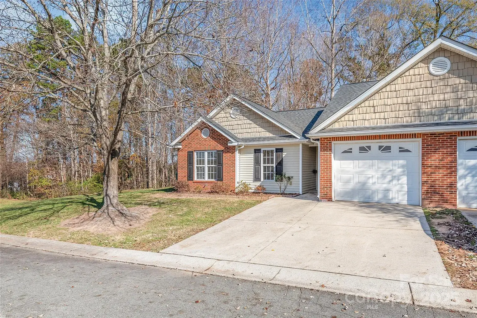 Front exterior of a single-story brick-and-siding home with attached garage, driveway, and wooded backdrop in Mount Holly, North Carolina.