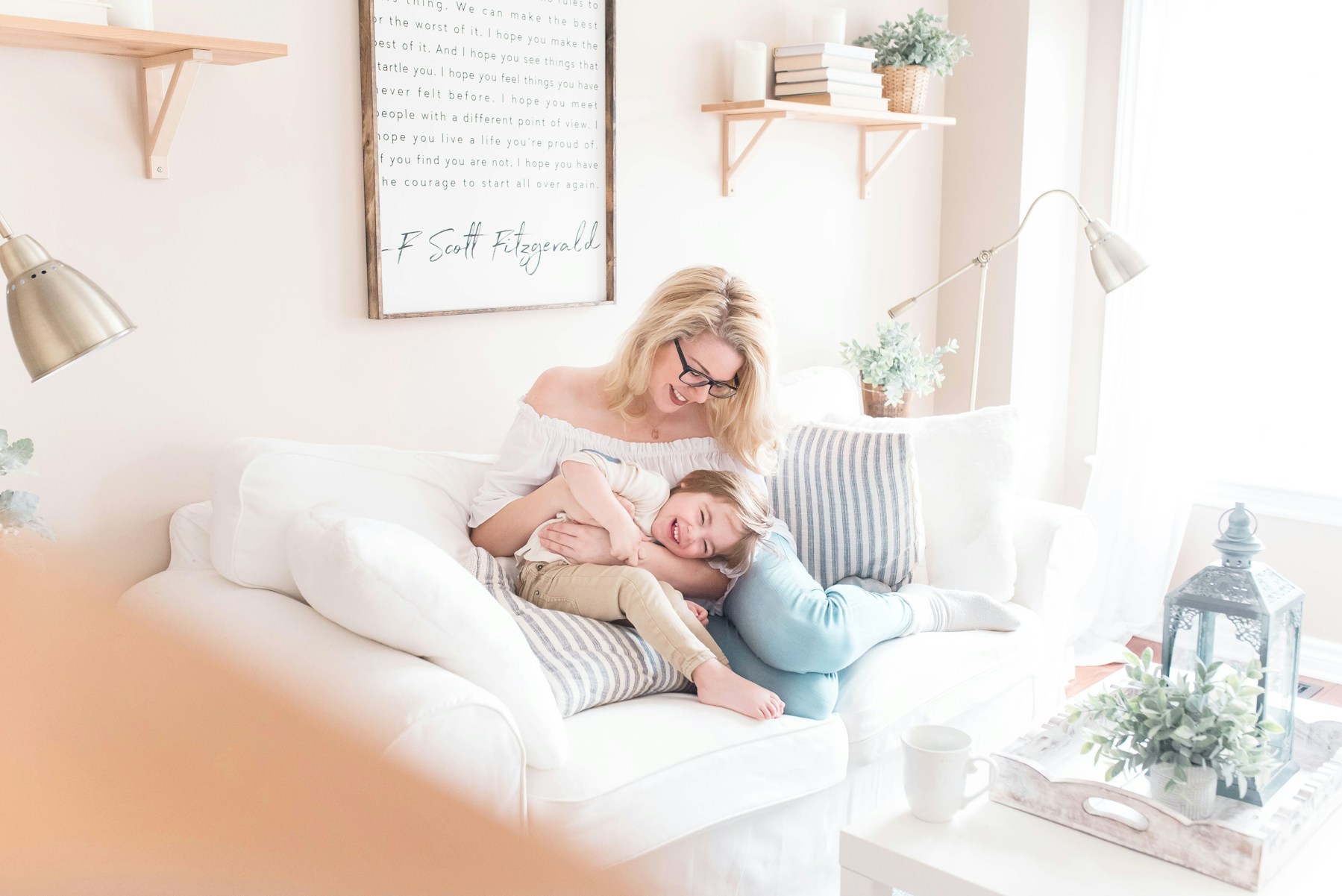 Bright living room with a mother hugging her young child on a white sofa, surrounded by cozy décor, striped pillows, and natural light.